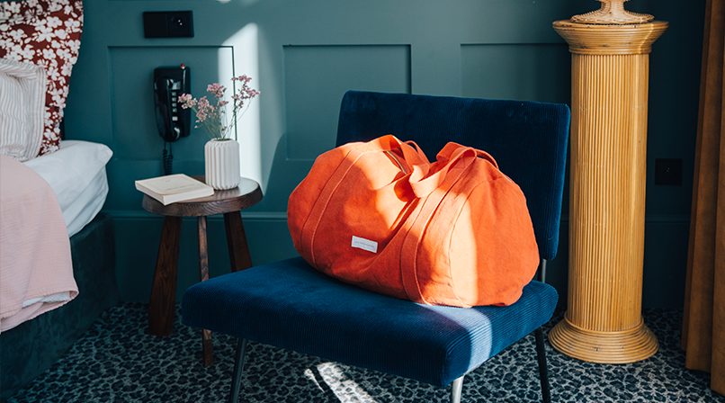 Une femme portant un grand sac cabas orange vif marche en extérieur, éclairée par le soleil, près d'un mur en pierre et d'une porte noire.