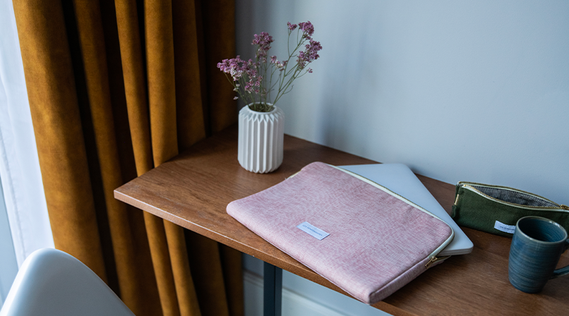 Un bureau en bois avec une housse d'ordinateur rose, un vase blanc avec des fleurs séchées, un sac vert et une tasse en céramique bleue, le tout encadré par des rideaux moutarde.